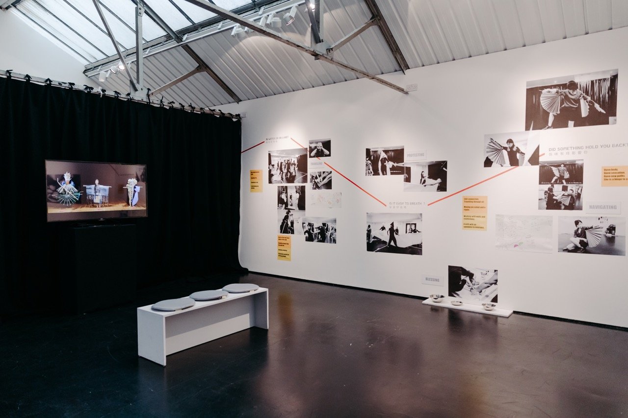 a photo of the exhibition space shows a video screen on a black curatined wall on the left with a mythical person performance and on the right with monochrome photos mindmapped across the white wall