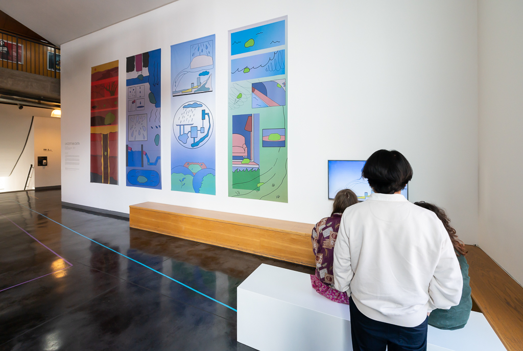 an exhibibition space with four colourful comic strip banners hang on a tall wall as 3 people view a video monitor to the right